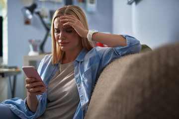 Upset young woman in blue shirt using cellphone at home