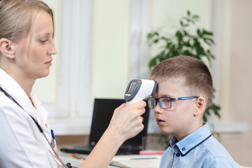 Dłoń lekarki mierzy temperaturę chłopcu w niebieskiej koszuli i okularach, za pomocą elektronicznego termometru na podczerwień. - obrazy, fototapety, plakaty