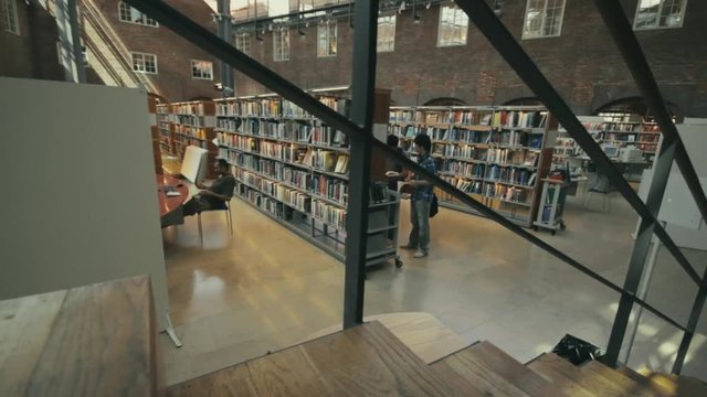 Students study inside a bright and spacious university library. Two young men stand at a long row of bookshelves selecting a book for their research. Camera descending stairs behind railing.