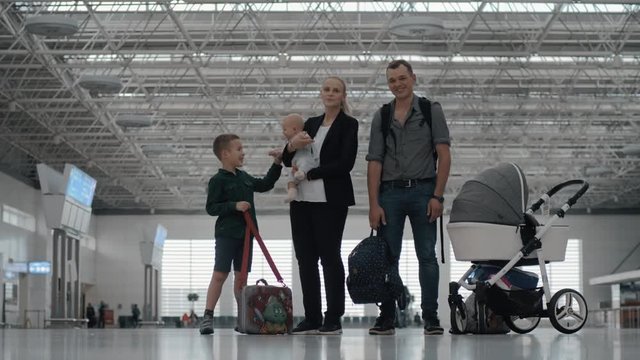 Happy family of four at the airport. Cheerful parents and children waiting for the flight. Elder son playing with baby in mothers hands