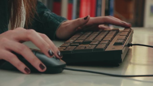 Close up of woman with long dark hair wearing a denim jacket using computer. Focus on hands with dark nail polish on the mouse and keyboard. Girl clicking computer mouse and typing on keyboard.
