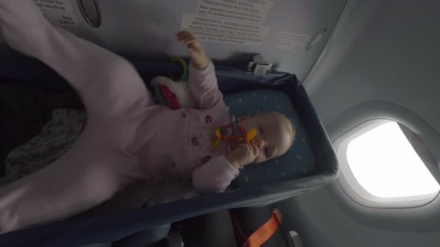 Active eight months baby girl lying in bassinet and playing with toy in the airplane. Comfortable air travel with little children
