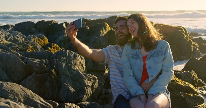 Smiling happy young couple clicking selfie on rock at the beach 4k