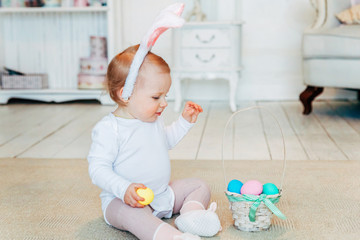 Fototapeta na wymiar Little child girl wearing bunny ears on Easter day. Girl holding basket with painted eggs sitting on floor at home, having fun on Easter egg hunt. Happy Easter holiday celebration spring concept