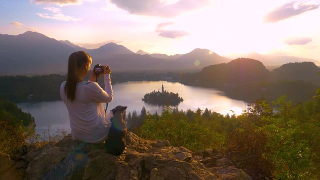 SLOW MOTION, LENS FLARE, CLOSE UP, COPY SPACE: Traveler girl sitting next to her cute puppy and taking photos of golden summer sunset illuminating the famous church in the middle of tranquil lake Bled