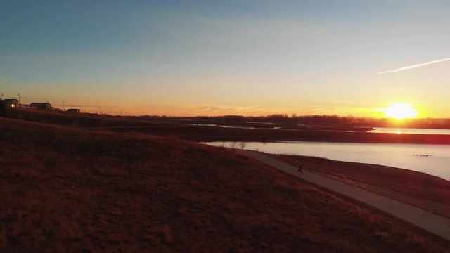 Electric Skateboarding on Curvy Path with Lake and Sun at Sunrise Aerial Leading View
