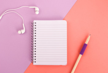 Top view of spiral notepad, wooden pencil and headphones on pastel purple and coral background. Flat lay style. Place for text.