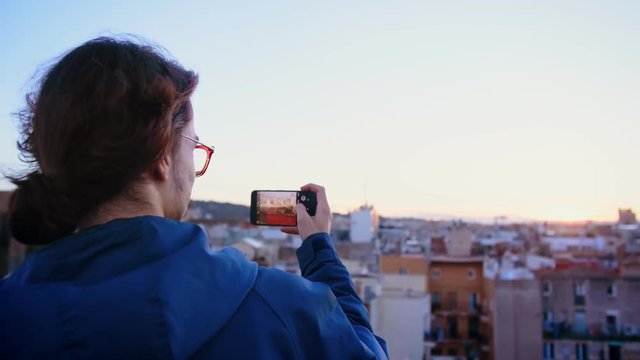 Young ambitious hipster student in new in city want to capture important moment and making smartphone or mobile phone camera photo or picture of view of rooftop during sunset or sunrise in Europe town