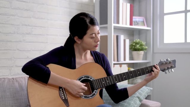 Pretty young asian woman playing guitar while sitting on sofa in light living room. happy girl practicing instrument for band performing. female player love music lifestyle concept.