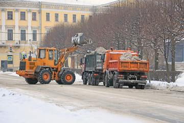 a tractor removing snow from the street into big trucks on a winter city street