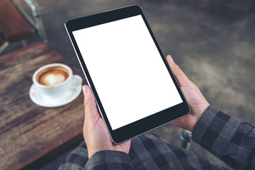 Mockup image of woman's hands holding black tablet pc with blank screen with coffee cup on wooden table in cafe