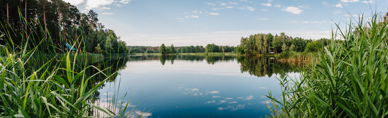 Fototapeta na wymiar Tranquil landscape at a lake, with the vibrant blue sky, white clouds and the trees reflected symmetrically in the clean blue water. Summer. Lake with spring trees panorama photo.