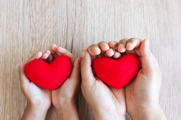 Fototapeta premium Red heart in child hands and parent hands on wooden table background with love and harmony