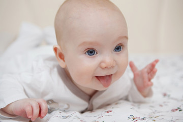 Beautiful five-month blue-eyed baby girl lies on her stomach. Shows tongue, various emotions and a smile on the face. Infant look. Maternal care. Childcare. Close-up.