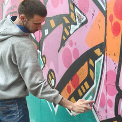 A young guy in a gray hoodie paints graffiti in pink and green c