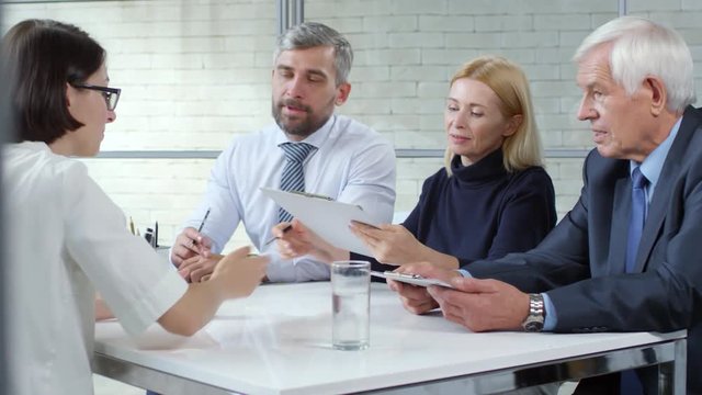 Tracking shot of bearded businessman, blond businesswoman and elderly manager in formalwear conducting job interview with young woman in glasses. They are reviewing her CV and having conversation
