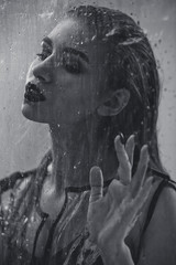 Black and white shot of model with wet hair dressed in black swimming suit and transparent rain coat is posing behind the wet transparent membrane 