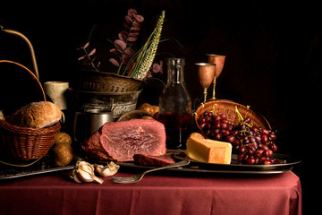Fine art food image reminiscent of 17th Century still life painting featuring meat, cheese, grapes, bread and garlic. Shot with various props relevant to that era and lit with moody studio flash