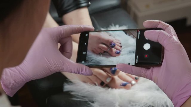 The pedicure master in pink gloves holds the phone and takes pictures of the client's painted and built-up nails with the help of the camera on the phone. Concept of beauty and health