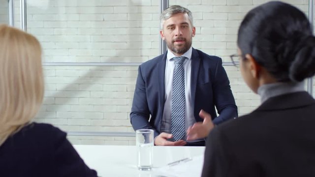 Tracking shot of bearded man in suit and necktie handing his CV to businesswomen conducting job interview, then introducing himself and answering questions about his professional experience