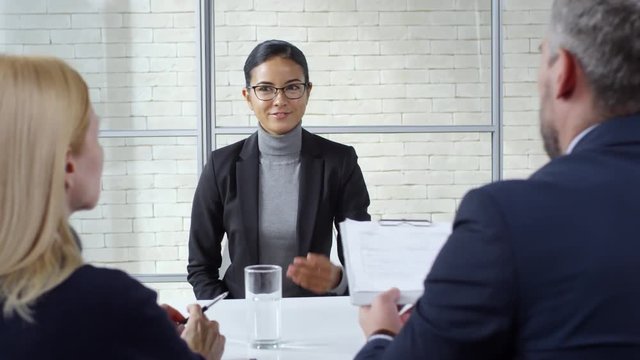 Tracking shot of young woman in glasses handing her CV to businessman and businesswoman conducting panel job interview. She is smiling and introducing herself
