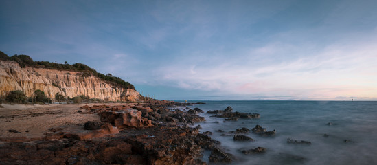 Fototapeta na wymiar Panoramic Long Exposure Shot of Red Bluff Lookout, a burnt orange-colour cliff, characterised by the oxidised iron in the cliffs with a rocky beach in the sunset time