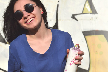 Portrait of an emotional young girl with black hair and piercings. Photo of a girl with aerosol paint cans in hands on a graffiti wall background. The concept of street art and use of aerosol paints
