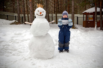 A little boy in the winter in the garden stands next to a snowman and looks at the camera smiling