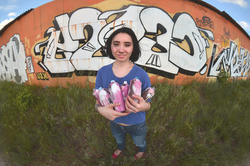 Portrait of an emotional young girl with black hair and piercings. A wide-angle photo of a girl with aerosol paint cans in the hands on a graffiti wall background. A modern portrait of a fisheye lens