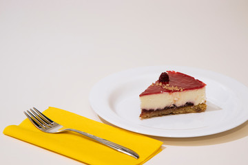 a slice of cheesecake on a white plate, with yellow napkin and steel cake fork. White background.