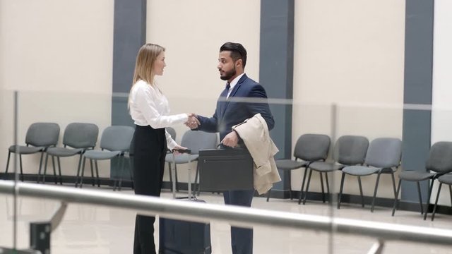 Cross-section shot of Caucasian businesswoman with rolling-bag meeting Arabian businessman in lobby of building, shaking hands and following him