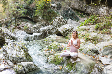 Relaxed beautiful blonde woman yoga in a bodycon white suit meditates sitting in a lotus position on a stone in the middle of beautiful wildlife with a mountain stream. Nature's inspiration concept