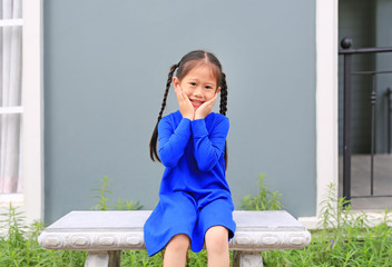 Cheerful little child girl sitting on stone bench with touching her cheek and smile.