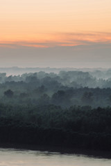 Mystical view on riverbank  of large island with forest under haze at early morning. Eerie mist among layers from tree silhouettes under predawn sky. Morning atmospheric landscape of majestic nature.