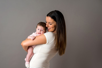 A beautiful young mother standing and holding her baby girl in her arms in front of a grey background in a studio.