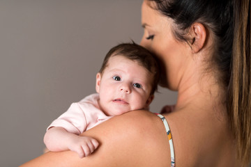 Closeup photo of a baby while her mother holding her on her chest while standing in front of a grey background in a studio.