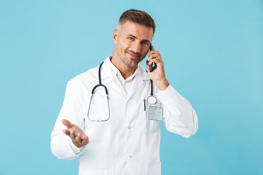 Photo of happy medical doctor wearing white coat and stethoscope talking on cell phone, standing isolated over blue background