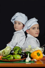 The boys in the cooks costumes cut the salad in the kitchen. Children prepare healthy food. Friendly cooperation of cooks. Isolated on black background.