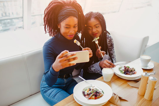 Two african models sit and pose on phone camera. They hold forks with food close to mouth. People are in cafe.
