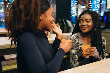 Cheerful african women sit in cafe and talk. They smile to each other. Models keep glasses with juice in hands.