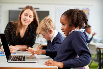 A female school teacher sitting at a desk helping a boy and girl using a laptop computer in primary...