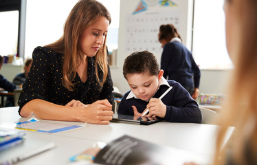 Young female teacher working with a Down syndrome schoolboy sitting at desk using a tablet computer and stylus in a primary school classroom, selective focus