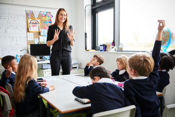 Female primary school teacher standing in a classroom gesturing to  schoolchildren sitting at a...