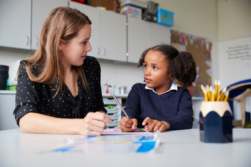 Young female primary school teacher working one on one with a schoolgirl at a table in a classroom, looking at each other, close up