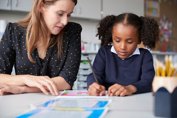 Young female primary school teacher working one on one with a schoolgirl watching her write at a...