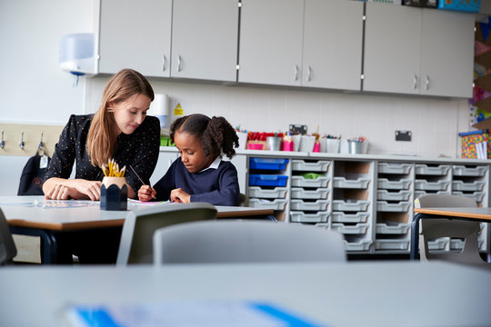 Young Female Primary School Teacher Working One On One With A Schoolgirl Sitting At Table In A Classroom, Selective Focus