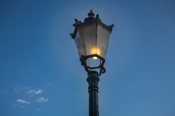 Fototapeta na wymiar Outdoor Victorian Style Pillar Lantern. Roadside Black Iron Cast Single Lamp Post. Streetlight with Dirty Smoked Glass and Lighted Bulb against Blue Sky