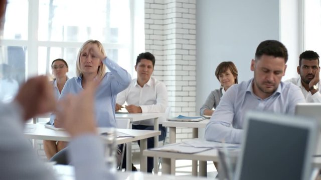 Diverse group of business people sitting in seminar room and listening to female coach during corporate training; blonde businesswoman putting her hand up and asking a question