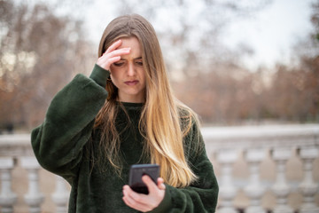 Troubled young woman rubbing forehead and looking at modern smartphone while standing in autumn park
