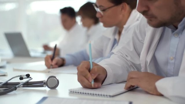 Rack focused shot of group of healthcare specialists sitting at desk and writing down in notebooks while taking a test during medical training course